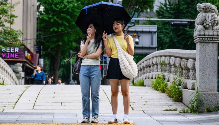 Pedestrians use an umbrella to shield from the sun and a portable fan as they wait to cross a road in Seoul on August 14, 2024, during a prolonged heatwave which has gripped much of the country. — AFP