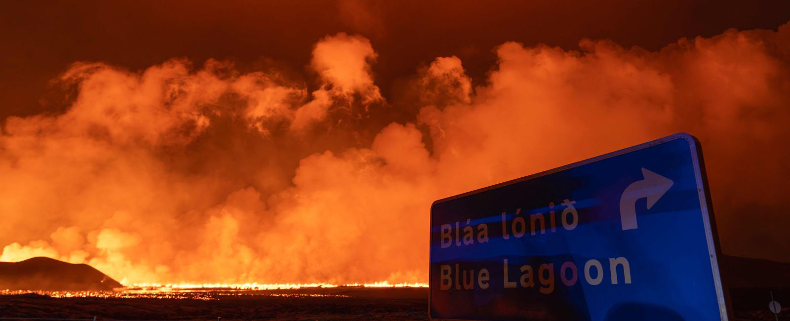 Street sign showing the way to the Blue Lagoon with the lava pouring and spreading from the volcanic eruption in the background near the Grindavik on the Icelandic peninsula of Reykjanes on August 23, 2024. — AFP