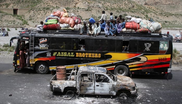 A bus with passengers sitting on the roof with belongings, drives past a damaged vehicle, a day after militants conducted deadly attacks, in Bolan district of Balochistan on August 27, 2024. Reuters