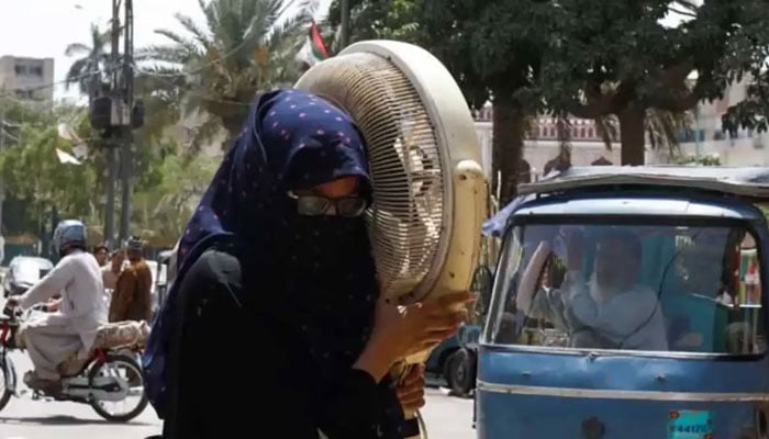 A woman carries a pedestal fan as she walks towards a repair shop during hot and humid weather in Karachi. — Reuters/File